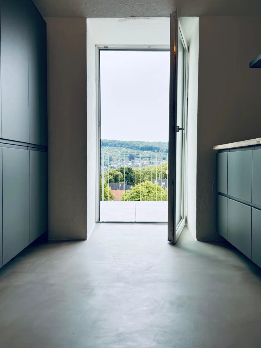 Minimalistischer Betonraum mit offener Balkontür, Einbauschränken und grüner Aussicht.