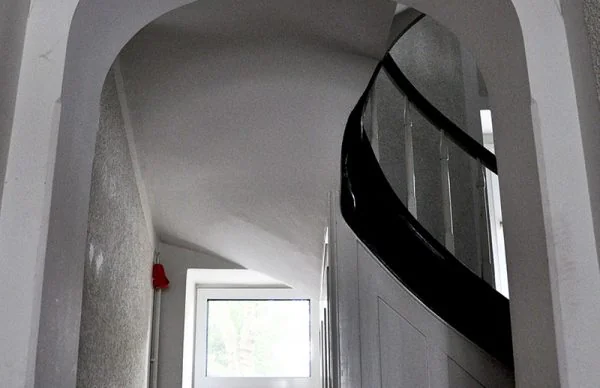Historisches Treppenhaus mit Rundbogen, geschwungener Holztreppe und hellem Fenster.