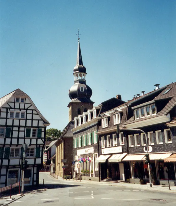 Historische Altstadt Cronenberg mit Kirchturm, Fachwerkhaus und Geschäften bei blauem Himmel.
