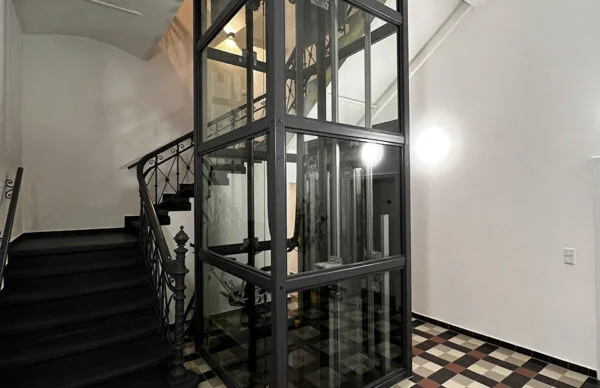 Glasaufzug neben historischem Treppenhaus mit schmiedeeisernem Geländer und Fliesenboden.