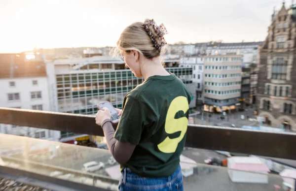 Person mit Smartphone blickt vom Greenhouse am Neumarkt auf die Kölner Innenstadt.