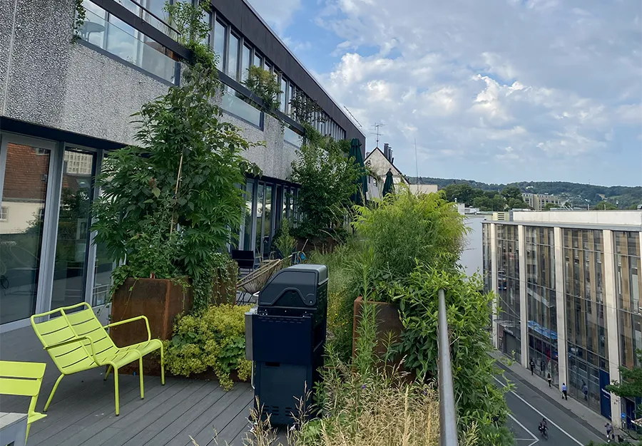 Begrünte Dachterrasse im vierten Stock mit Stadtblick, Betonfassade und modernen Gartenmöbeln.