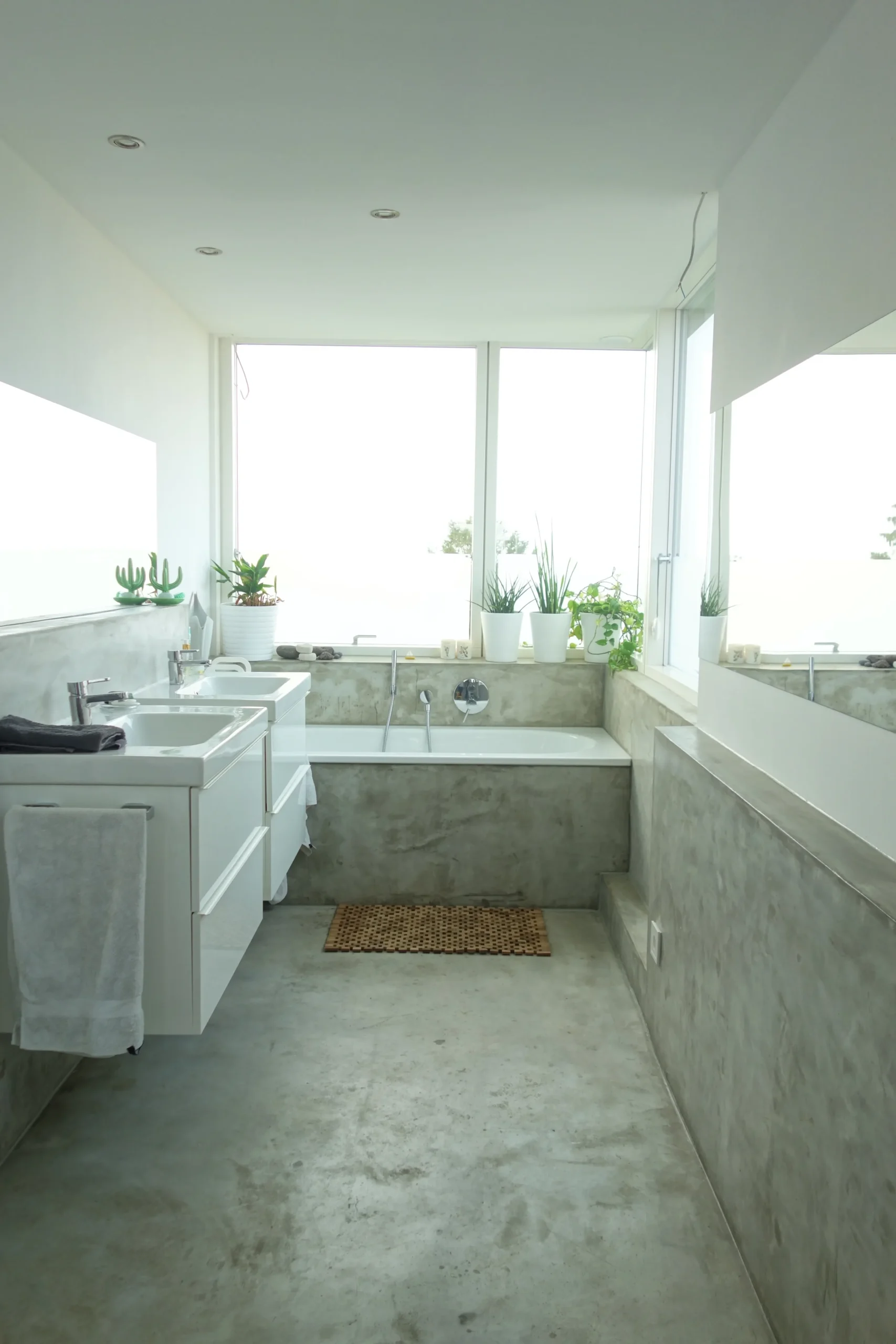Modernes Betonbad mit eingelassener Badewanne, Doppelwaschbecken, Pflanzen und Tageslicht.
