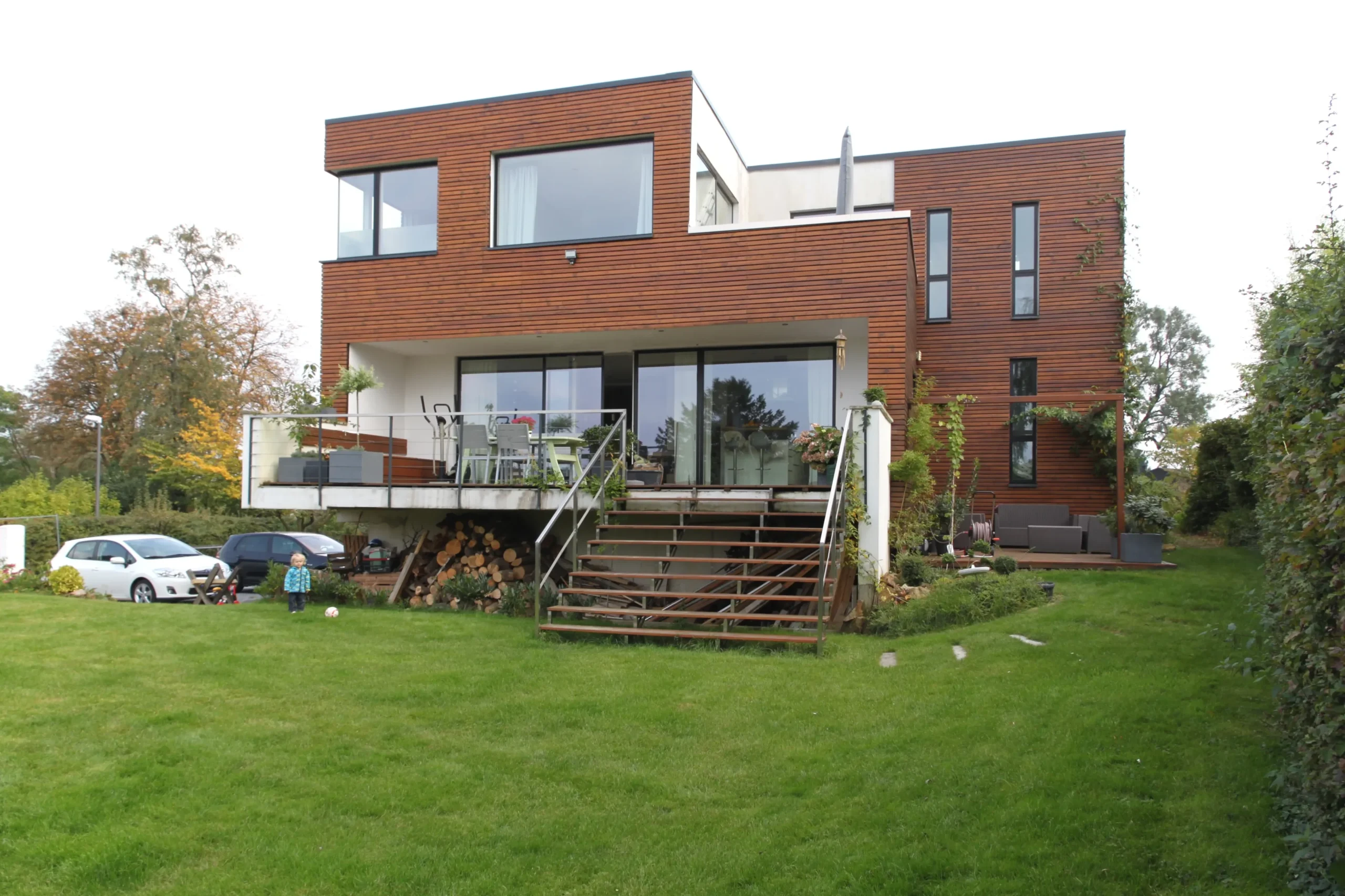 Modernes Holzhaus mit Garten, Terrasse, Balkon und Kind auf Rasen.