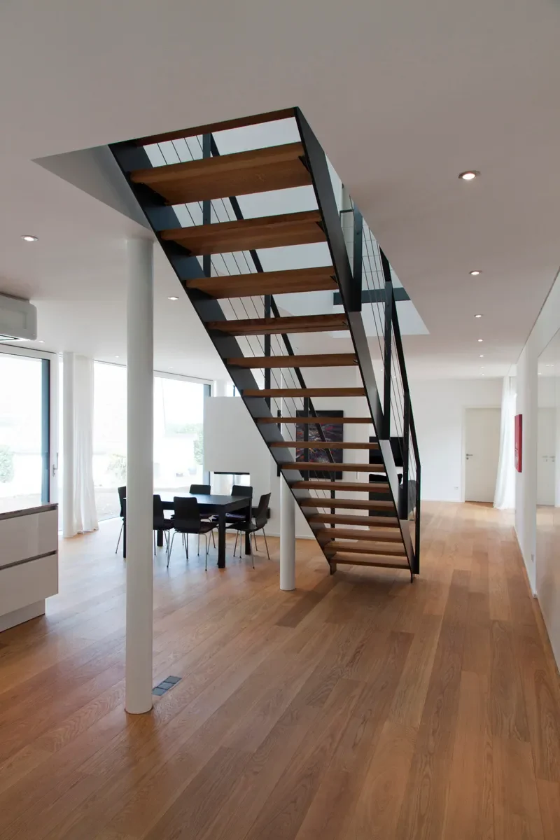 Modernes Hausinterieur mit freischwebender Holztreppe, offenem Wohnbereich und großen Fenstern.