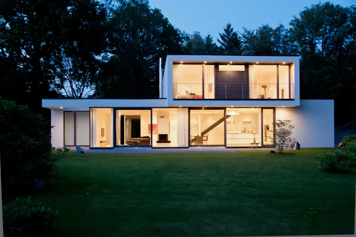 Modernes Einfamilienhaus mit Glasfassade, Garten und warm beleuchteter Architektur bei Dämmerung.