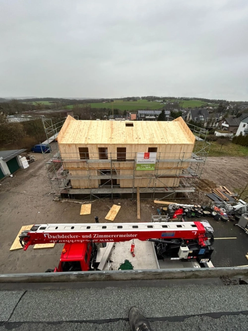 Holzhaus im Bau mit Kran, Dachdecker und Gerüst auf ländlicher Baustelle.