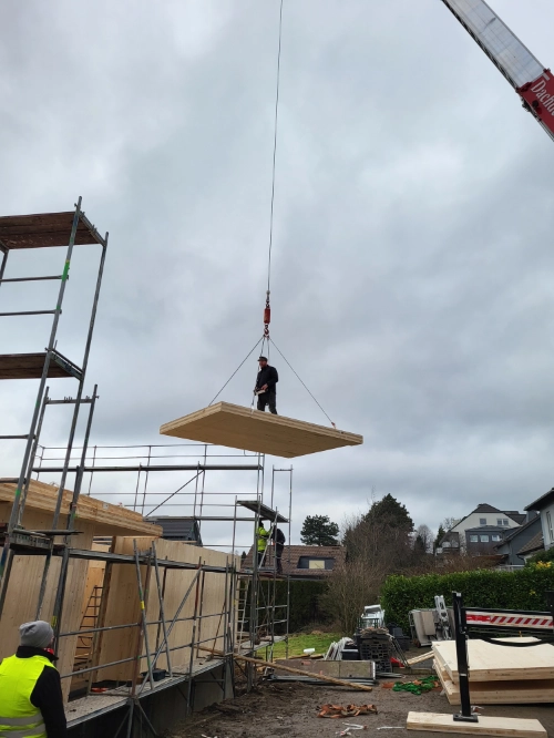 Holzhaus-Baustelle mit Kran, Arbeiter und schwebendem Holz-Fertigelement.
