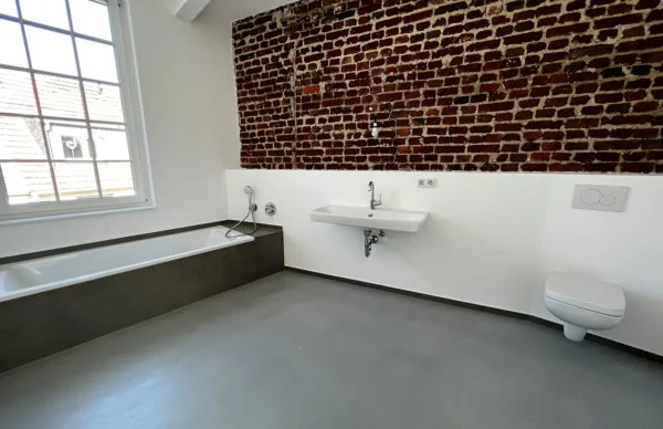 Modernes Loft-Badezimmer mit Backsteinwand, eingebauter Badewanne, Waschbecken und viel Tageslicht.