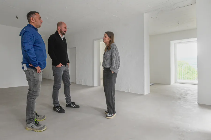 Drei Menschen besichtigen einen hellen Bunker-Rohbau mit Betonboden und weißen Wänden.