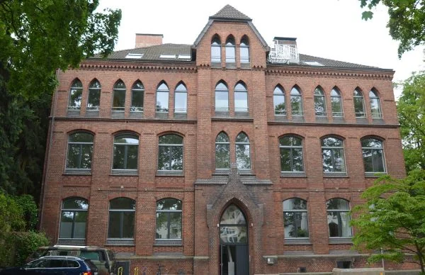 Historisches Backstein-Schulgebäude mit saniertem Eingang und gotischen Fenstern im Grünen.