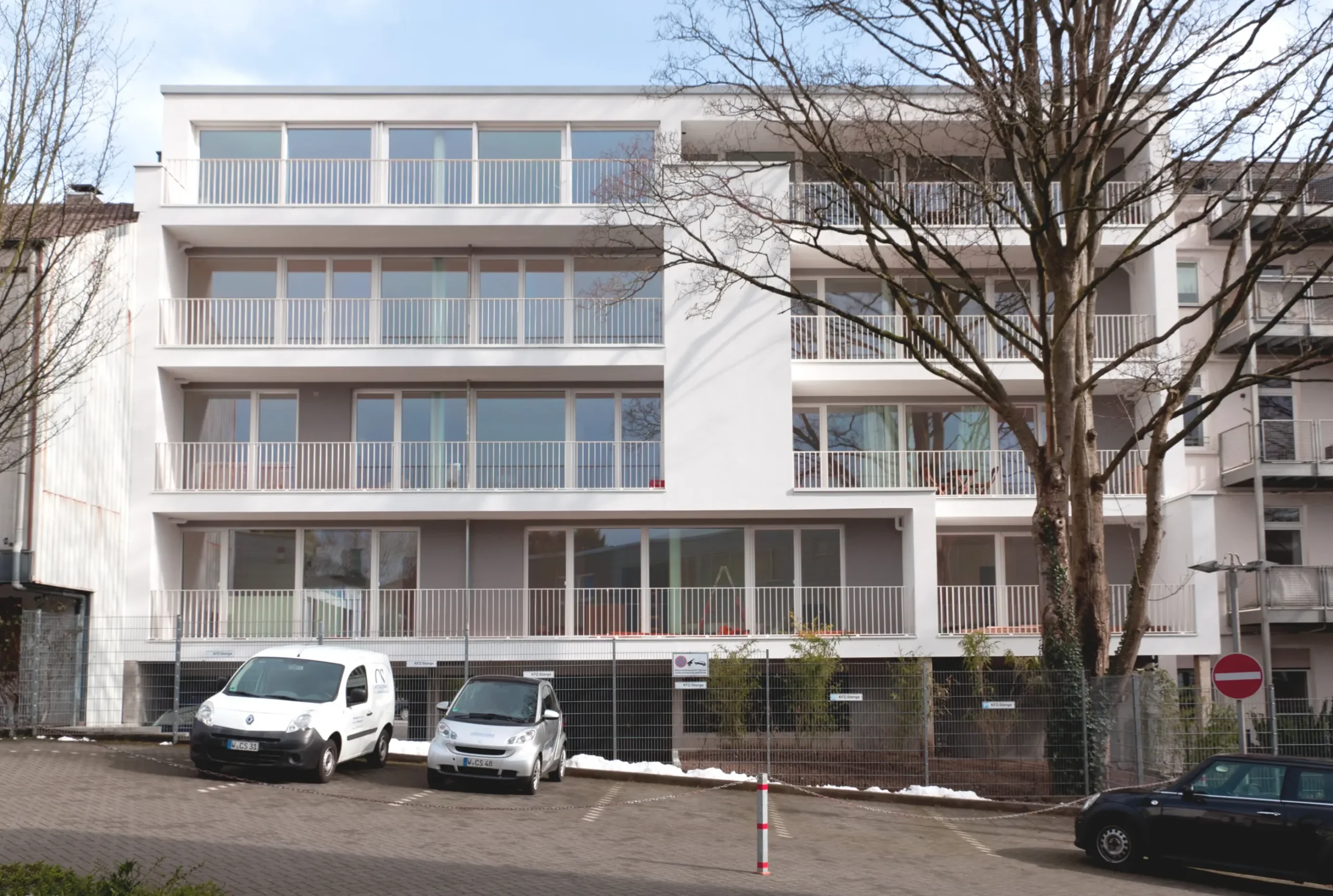Modernes Mehrfamilienhaus mit Balkonen, Glasfassade, Parkplatz und Schnee in urbaner Wohnanlage.