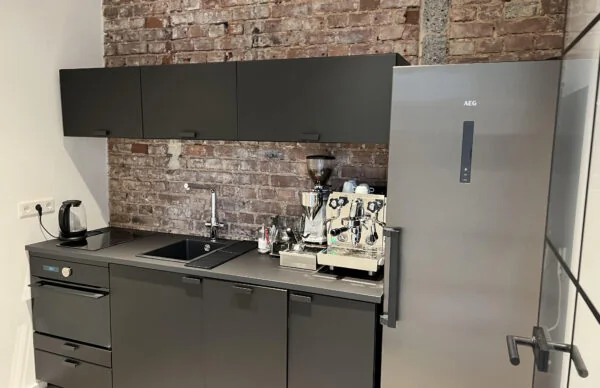 Moderne schwarze Büroküche mit Backsteinwand, Edelstahlkühlschrank und professioneller Kaffeemaschine.