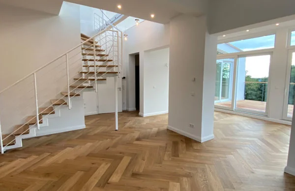 Heller offener Wohnraum mit Fischgrätparkett, moderner Treppe und bodentiefen Fenstern.