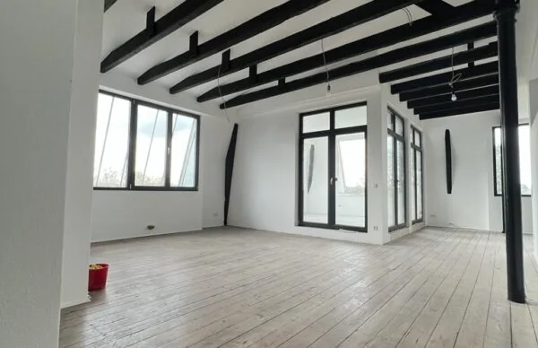 Helles Loft mit Holzboden, schwarzen Deckenbalken und großen Fenstern.