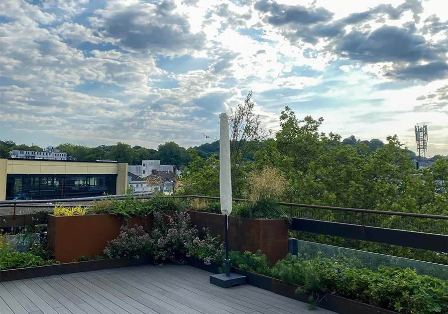 Dachterrasse mit Dachgarten, Sonnenschirm und Skyline-Ausblick über grüne Stadtlandschaft.